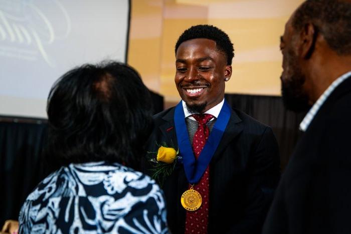 Award recipient Kader Kohou faces the camera and smiles with eyes downcast and a gold medal around his neck. Two people stand in front of him with their backs to the camera.